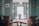 A vintage-style room with teal walls and wooden chairs set around a small round table. The table holds a decorative vase with flowers. The walls are adorned with framed photographs and stained glass panels. Ambient light from a globe ceiling lamp softly illuminates the space, lending it a cozy, nostalgic feel.
