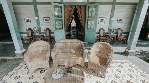 A patio setting with wicker furniture, including two chairs and a sofa, arranged on a decorative tiled floor. Behind the seating area are two green wooden doors with stained glass, slightly ajar, revealing a glimpse inside the room. The interior wall is adorned with framed pictures and vintage-style chairs placed against it. There are small tables with potted plants next to the chairs.