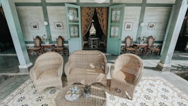 A patio setting with wicker furniture, including two chairs and a sofa, arranged on a decorative tiled floor. Behind the seating area are two green wooden doors with stained glass, slightly ajar, revealing a glimpse inside the room. The interior wall is adorned with framed pictures and vintage-style chairs placed against it. There are small tables with potted plants next to the chairs.