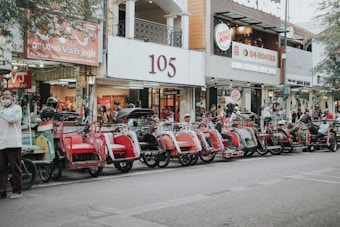A row of traditional rickshaws parked along a street in front of various shops. The rickshaws feature vibrant colors like red and green, and drivers are seen sitting or standing nearby. The area appears to be a busy urban location with signs for businesses visible in the background, including a Burger King.