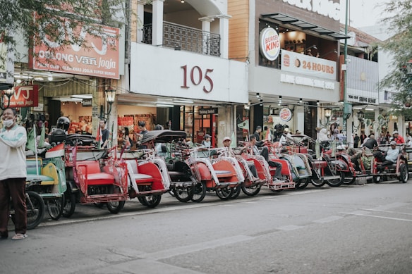 A row of traditional rickshaws parked along a street in front of various shops. The rickshaws feature vibrant colors like red and green, and drivers are seen sitting or standing nearby. The area appears to be a busy urban location with signs for businesses visible in the background, including a Burger King.