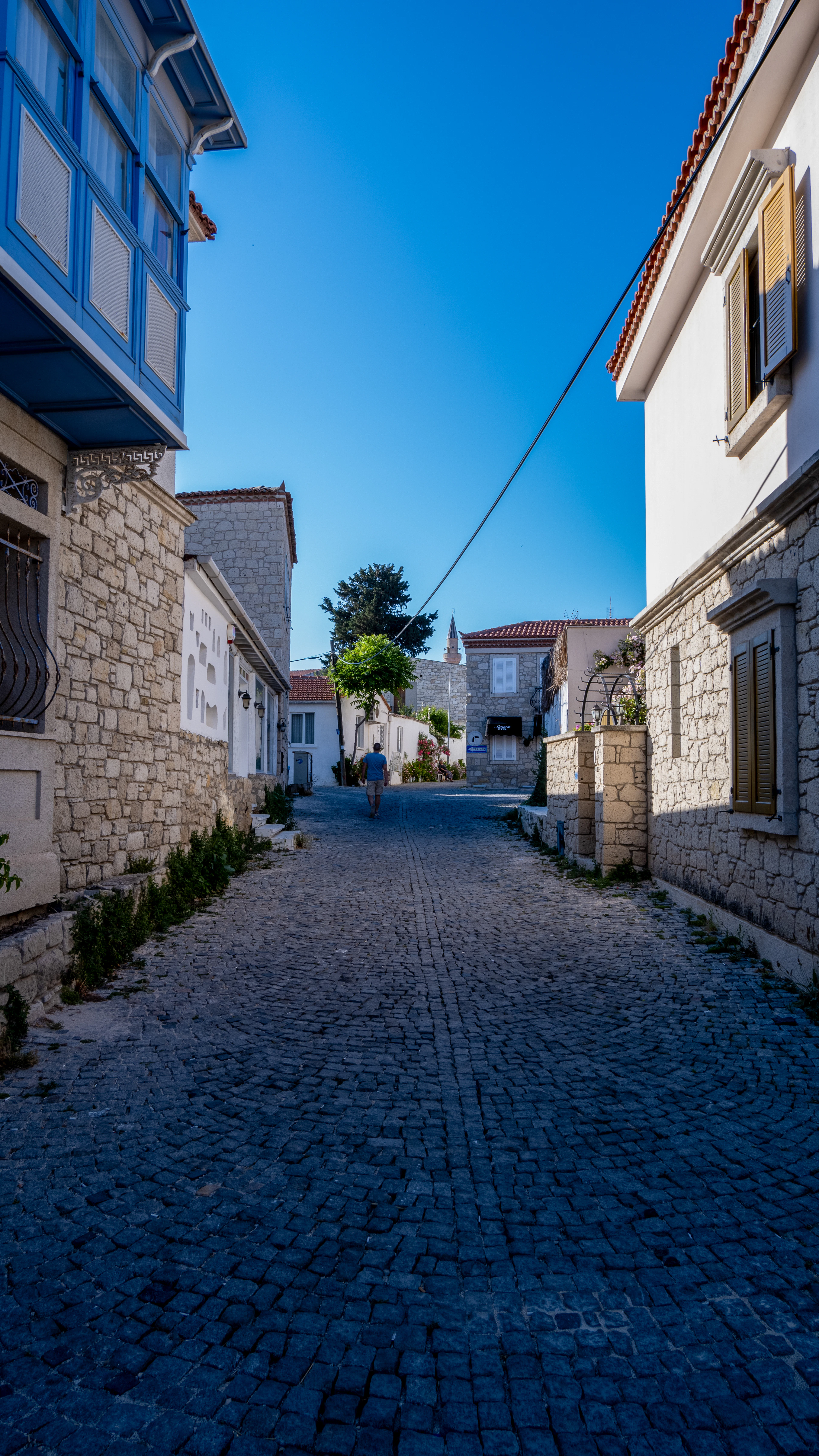 An empty cobbled street in Alaçatı between stone houses with blue shutters, with a lone walker in the distance