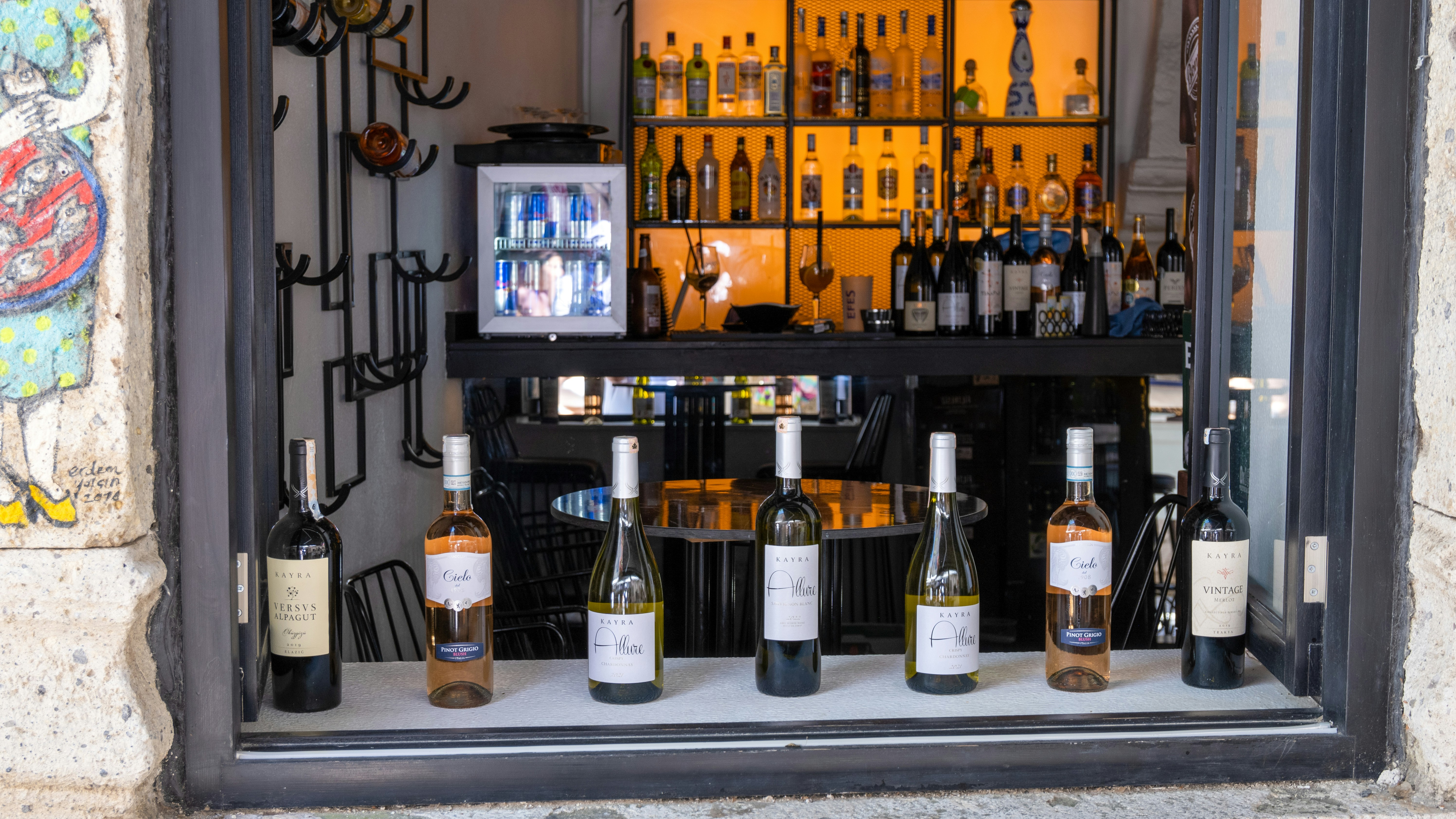 a bunch of bottles of wine sitting in a window, Alaçatıda hem yerel hem dünya çapında çok güzel içkiler bulabilirsiniz. You can find very good local and international drinks in Alaçatı.