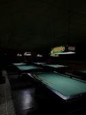 A dimly lit room containing several pool tables with green felt surfaces. Overhead, illuminated signs are visible, displaying the brand name 'Stag Lager Beer' in bright colors. The room has a gloomy atmosphere due to the low lighting conditions, with shadowed areas around the tables.