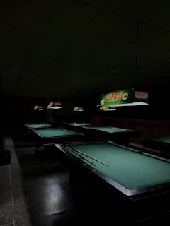 A dimly lit room containing several pool tables with green felt surfaces. Overhead, illuminated signs are visible, displaying the brand name 'Stag Lager Beer' in bright colors. The room has a gloomy atmosphere due to the low lighting conditions, with shadowed areas around the tables.