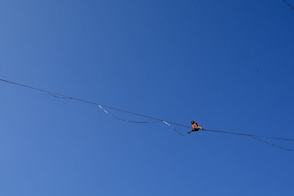 A person is performing a balance act on a tightrope against a clear blue sky. The individual is positioned towards the right side of the image, focusing intently on maintaining balance.