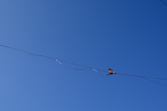 A person is performing a balance act on a tightrope against a clear blue sky. The individual is positioned towards the right side of the image, focusing intently on maintaining balance.
