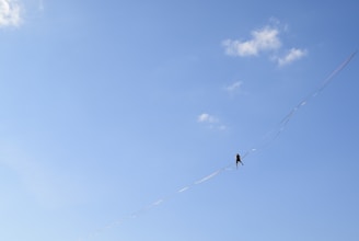 A person is walking on a high wire against a clear blue sky with a few scattered clouds. The wire stretches diagonally across the image, and the person appears small against the vast sky.