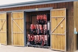 A wooden storage shed with two doors open, revealing neatly organized rows of red life jackets hanging inside. The shed has a rustic appearance with a natural wood finish, and sailing ropes are hanging from hooks on the door.