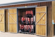 A wooden storage shed with two doors open, revealing neatly organized rows of red life jackets hanging inside. The shed has a rustic appearance with a natural wood finish, and sailing ropes are hanging from hooks on the door.