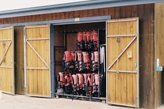 A wooden storage shed with two doors open, revealing neatly organized rows of red life jackets hanging inside. The shed has a rustic appearance with a natural wood finish, and sailing ropes are hanging from hooks on the door.