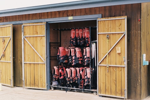 A wooden storage shed with two doors open, revealing neatly organized rows of red life jackets hanging inside. The shed has a rustic appearance with a natural wood finish, and sailing ropes are hanging from hooks on the door.