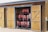 A wooden storage shed with two doors open, revealing neatly organized rows of red life jackets hanging inside. The shed has a rustic appearance with a natural wood finish, and sailing ropes are hanging from hooks on the door.