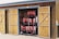 A wooden storage shed with two doors open, revealing neatly organized rows of red life jackets hanging inside. The shed has a rustic appearance with a natural wood finish, and sailing ropes are hanging from hooks on the door.