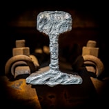 A close-up view of a metallic object resembling a railroad track section, with a rough, textured surface and a silvery sheen. The object is flanked by rusty bolts and fasteners, with a dark background emphasizing the industrial nature of the scene.