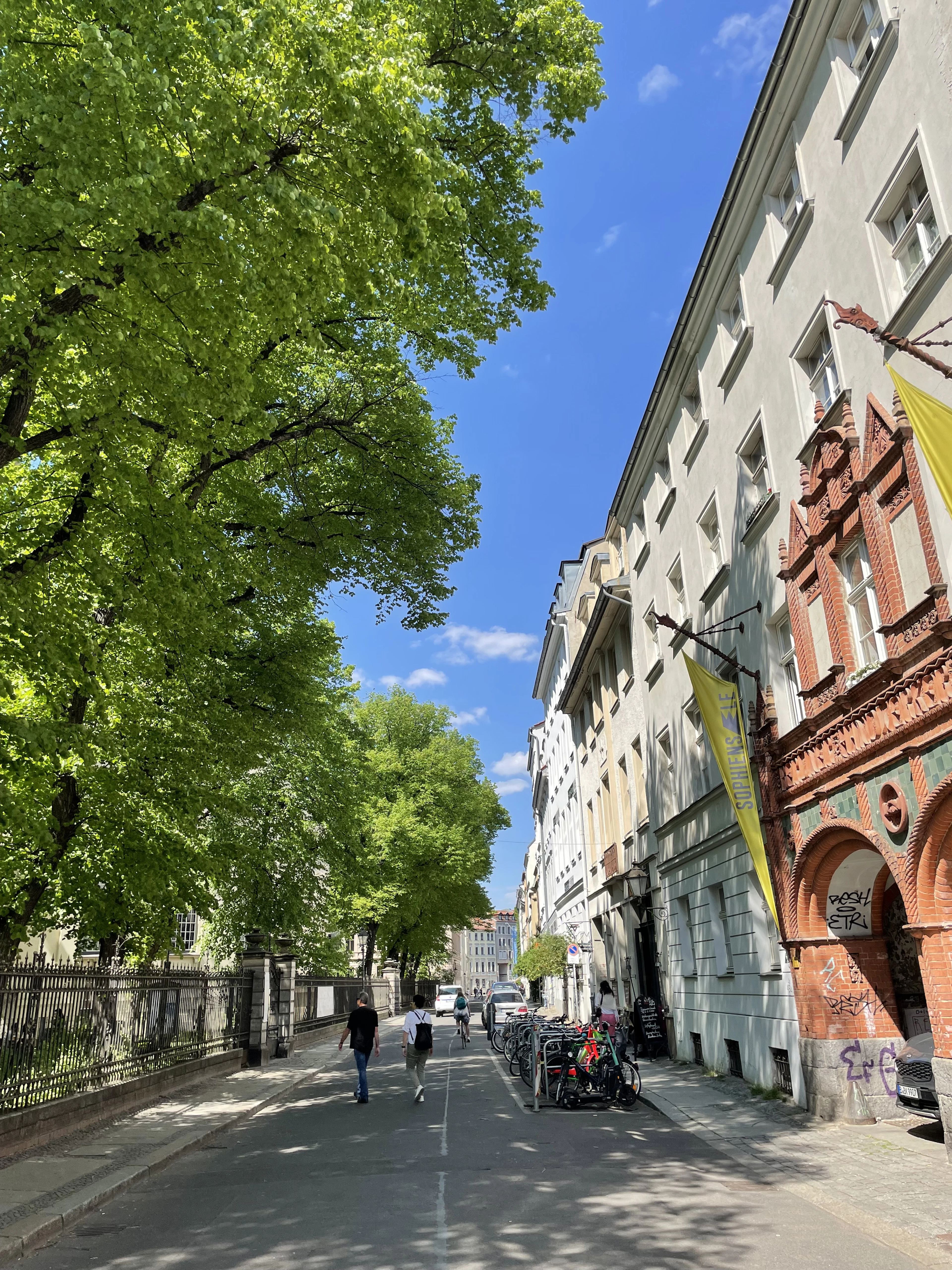 A quiet tree-shaded street in Berlin-Mitte on a summer morning — pale Altbau facades and an ornate red-brick gable on one side, racked bicycles at the kerb and two people walking down the middle.