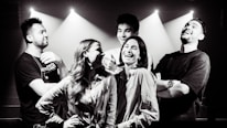 A group of five people stands together in an indoor setting with beams of light shining down, creating a dramatic effect. They appear to be enjoying each other's company, laughing and smiling, indicating a lively and cheerful interaction.