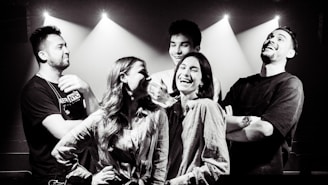 A group of five people stands together in an indoor setting with beams of light shining down, creating a dramatic effect. They appear to be enjoying each other's company, laughing and smiling, indicating a lively and cheerful interaction.