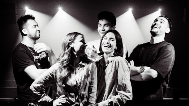A group of five people stands together in an indoor setting with beams of light shining down, creating a dramatic effect. They appear to be enjoying each other's company, laughing and smiling, indicating a lively and cheerful interaction.
