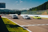 Three sports cars are racing on a track surrounded by lush green hills and trees. The sky is clear with a few clouds, and there is a fence along one side of the track. The cars are sleek and positioned on a wide, paved racing circuit under bright sunlight.