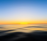 A serene ocean scene with a smooth water surface reflecting the soft colors of a sunset. The horizon line blends into a gradient sky transitioning from deep blue to warm yellow and orange hues, creating a calm and peaceful atmosphere.
