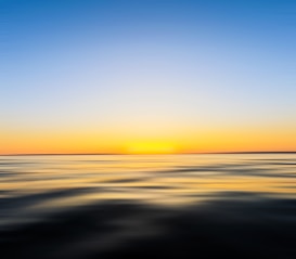 A serene ocean scene with a smooth water surface reflecting the soft colors of a sunset. The horizon line blends into a gradient sky transitioning from deep blue to warm yellow and orange hues, creating a calm and peaceful atmosphere.