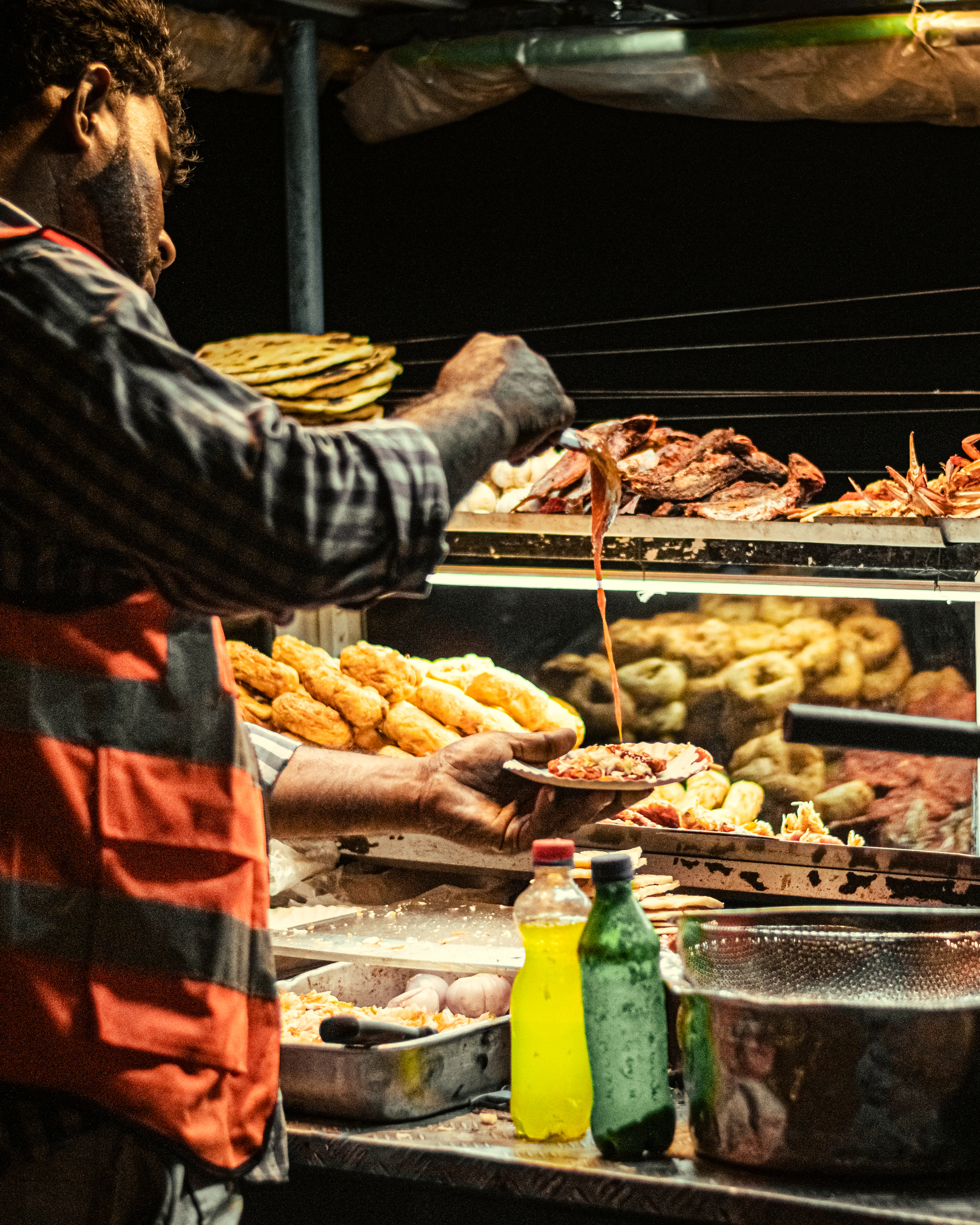 A Colombo street-food vendor pours sauce over a plate of isso wade at his night stall, the lit counter behind him stacked with golden prawn fritters, vadai, fried seafood and rolled roti, soda bottles glowing on the ledge