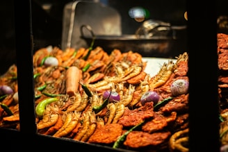 A display of various deep-fried foods, including golden-brown shrimp, red-colored patties, and whole onions. Green chilies are scattered throughout the arrangement, adding a splash of color. The scene is warmly lit and appears to be focused on street food or a market setting.