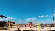 A beach volleyball match is taking place under a clear blue sky. Players in red and black outfits are actively engaged on the sandy court, with a volleyball mid-air. Spectators surround the court, watching the game. Colorful banners and advertisements are visible along the barriers.