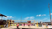 A beach volleyball match is taking place under a clear blue sky. Players in red and black outfits are actively engaged on the sandy court, with a volleyball mid-air. Spectators surround the court, watching the game. Colorful banners and advertisements are visible along the barriers.