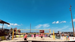 A beach volleyball match is taking place under a clear blue sky. Players in red and black outfits are actively engaged on the sandy court, with a volleyball mid-air. Spectators surround the court, watching the game. Colorful banners and advertisements are visible along the barriers.