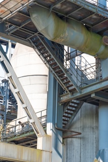 A complex structure of metal beams, staircases, and industrial pipes. The image features a large cylindrical tank and various metal platforms interconnected by stairs. The surfaces show signs of rust and wear, suggesting an outdoor industrial setting or factory.