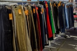 A variety of pants are neatly organized on a hanging rack, with different colors and styles visible. Price tags are attached to each pair, indicating they are for sale. The scene is set outdoors, with sunshine casting shadows on the ground.