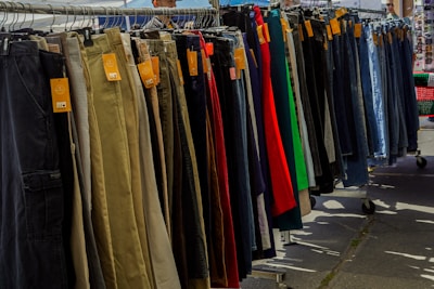 A variety of pants are neatly organized on a hanging rack, with different colors and styles visible. Price tags are attached to each pair, indicating they are for sale. The scene is set outdoors, with sunshine casting shadows on the ground.