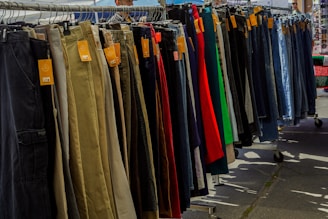 A variety of pants are neatly organized on a hanging rack, with different colors and styles visible. Price tags are attached to each pair, indicating they are for sale. The scene is set outdoors, with sunshine casting shadows on the ground.