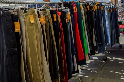 A variety of pants are neatly organized on a hanging rack, with different colors and styles visible. Price tags are attached to each pair, indicating they are for sale. The scene is set outdoors, with sunshine casting shadows on the ground.