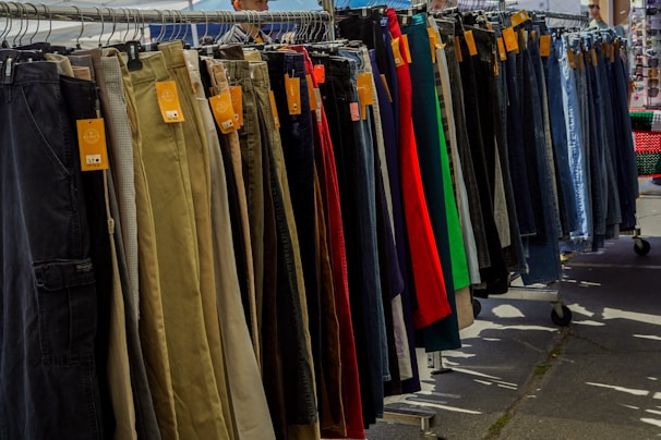 A variety of pants are neatly organized on a hanging rack, with different colors and styles visible. Price tags are attached to each pair, indicating they are for sale. The scene is set outdoors, with sunshine casting shadows on the ground.