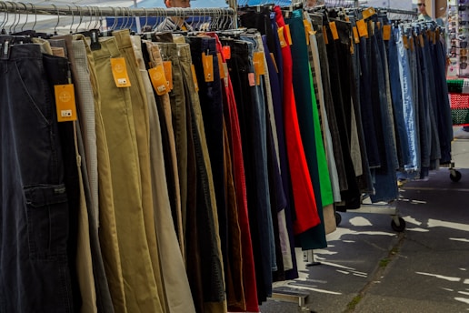A variety of pants are neatly organized on a hanging rack, with different colors and styles visible. Price tags are attached to each pair, indicating they are for sale. The scene is set outdoors, with sunshine casting shadows on the ground.