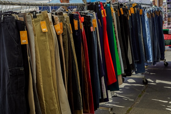 A variety of pants are neatly organized on a hanging rack, with different colors and styles visible. Price tags are attached to each pair, indicating they are for sale. The scene is set outdoors, with sunshine casting shadows on the ground.