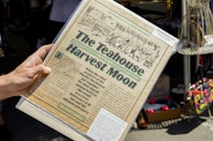 A hand is holding a vintage-style newspaper titled 'The Teahouse Harvest Moon.' The paper features an illustration of people and text describing an event blending aspects of a fair and a 'pot party.' It appears to be set in an outdoor market or fair with various items like shoes and colorful bags visible in the background.