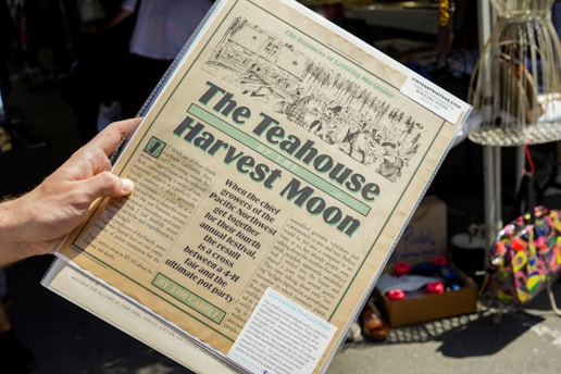 A hand is holding a vintage-style newspaper titled 'The Teahouse Harvest Moon.' The paper features an illustration of people and text describing an event blending aspects of a fair and a 'pot party.' It appears to be set in an outdoor market or fair with various items like shoes and colorful bags visible in the background.