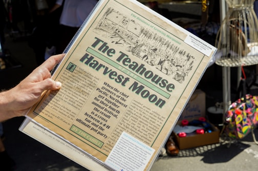 A hand is holding a vintage-style newspaper titled 'The Teahouse Harvest Moon.' The paper features an illustration of people and text describing an event blending aspects of a fair and a 'pot party.' It appears to be set in an outdoor market or fair with various items like shoes and colorful bags visible in the background.