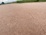A close-up view of a cracked concrete surface with a visible crack running through it. The background shows blurred green grass and a cloudy sky, indicating an outdoor setting.