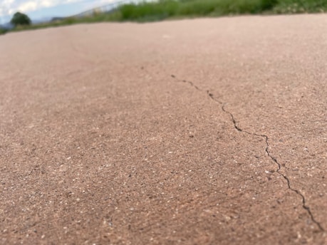 A close-up view of a cracked concrete surface with a visible crack running through it. The background shows blurred green grass and a cloudy sky, indicating an outdoor setting.