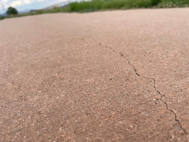 A close-up view of a cracked concrete surface with a visible crack running through it. The background shows blurred green grass and a cloudy sky, indicating an outdoor setting.