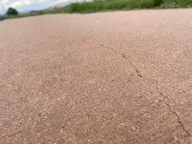 A close-up view of a cracked concrete surface with a visible crack running through it. The background shows blurred green grass and a cloudy sky, indicating an outdoor setting.