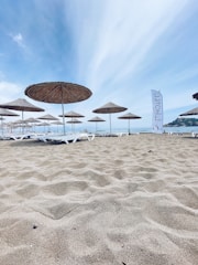 Several straw parasols are positioned on a sandy beach under a clear blue sky. White sun loungers are neatly arranged beneath the parasols, and a sign with the word 'Hotel' is visible on the right side. The shoreline is visible in the background, suggesting a calm seaside environment.
