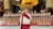 A person dressed in traditional robes stands with their back to the viewer, in front of an ornate altar filled with rows of offerings, colorful decorations, and statues. The setting appears to be a richly adorned place of worship, characterized by intricate patterns and vivid colors.