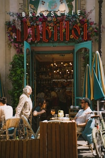 A lively street scene in front of a brasserie entrance adorned with vibrant flowers and neon signage. People are seated at outdoor tables, enjoying drinks while others are walking by. The atmosphere is bustling and warm, emphasized by the open doors revealing a glimpse of the cozy interior with a well-stocked bar.