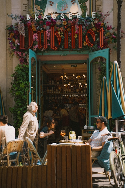A lively street scene in front of a brasserie entrance adorned with vibrant flowers and neon signage. People are seated at outdoor tables, enjoying drinks while others are walking by. The atmosphere is bustling and warm, emphasized by the open doors revealing a glimpse of the cozy interior with a well-stocked bar.